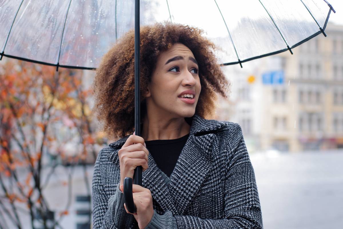 African American girl happy woman female tourist lady stands in strong wind rain storm cold windy autumn weather outside in city street with transparent umbrella laughing having fun looking into sky